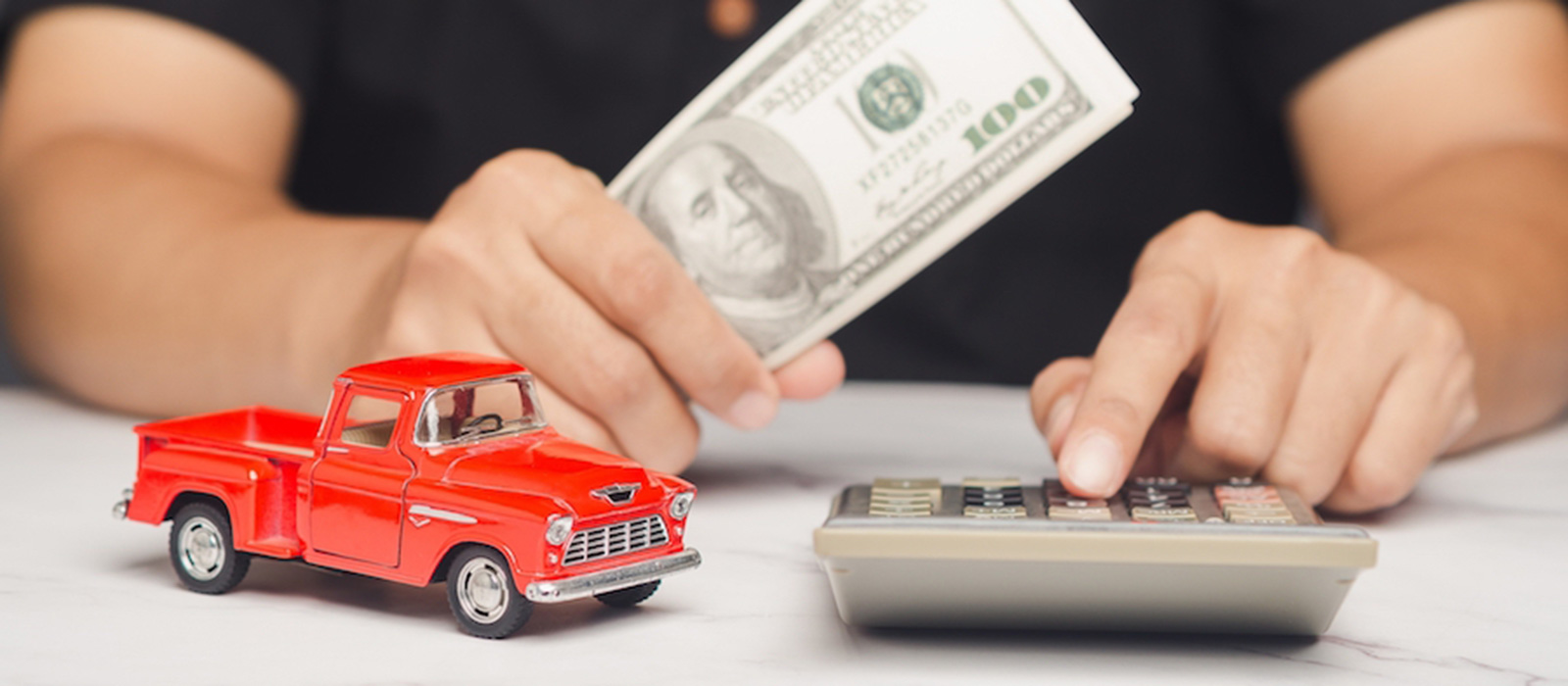 The torso and hands of a person with a calculator in one hand and a hundred dollar bill in the other, with a red toy pickup truck sitting on the table in front of them