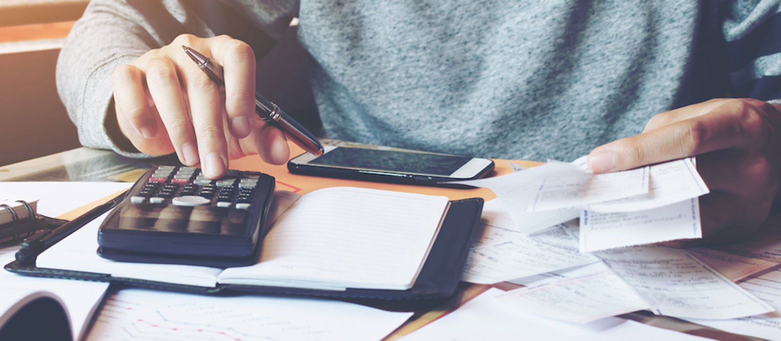 The hands and torso of a white person in a gray sweatshirt using a calculator at a table covered with papers