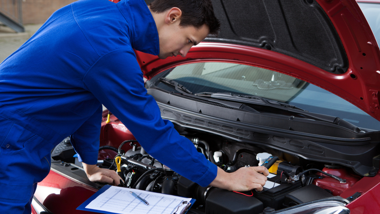 Mechanic in blue jumpsuit inspecting red car's engine with clipboard and diagnostic tool.