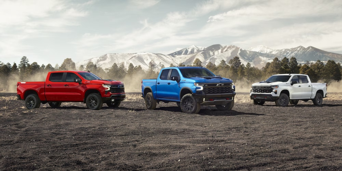 3 2026 Chevrolet Silverado pickup trucks, one red, one blue, and one white, parked on a dirt field with mountains in the background
