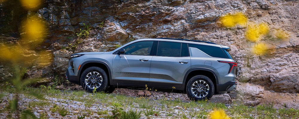 A side view of a gray 2026 Chevrolet Traverse parked in front of a rocky outcropping, with wildflowers in the foreground