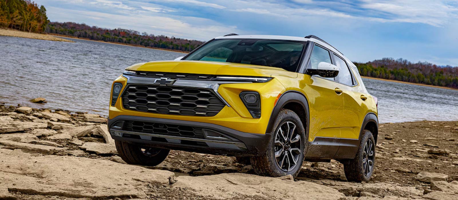 A yellow 2025 Chevrolet Trailblazer parked on the rocky shore of a lake